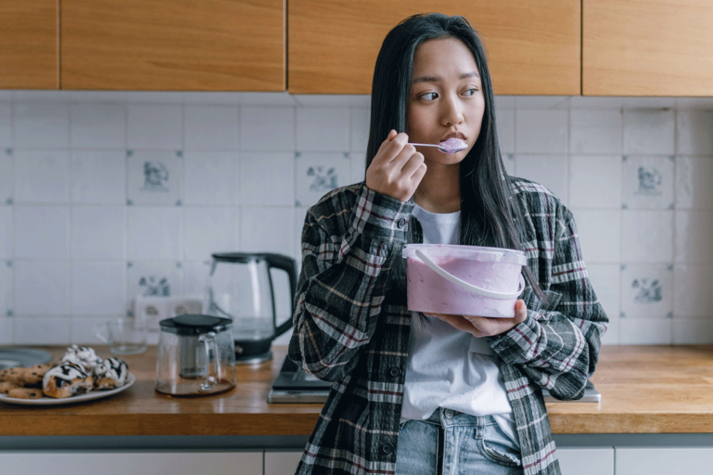 Girl standing in kitchen, eating yogurt and looking sad.