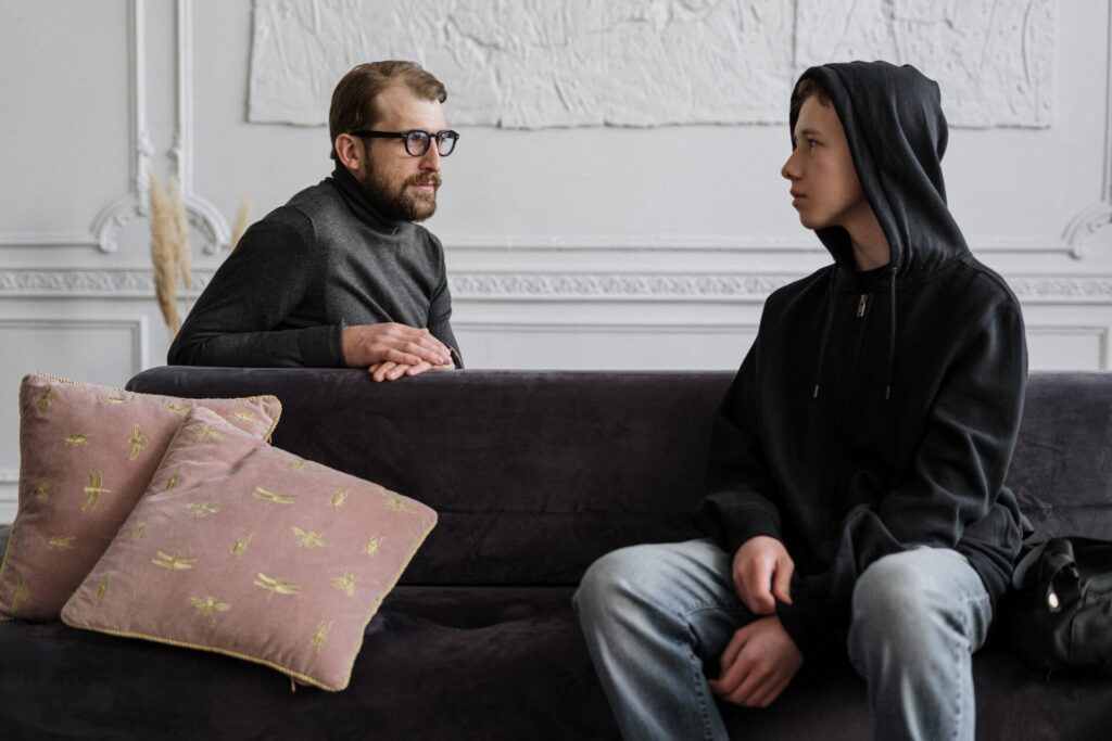 Boy in hoodie, sitting on couch and talking to older man.