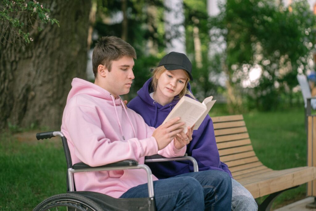 Man in wheelchair reading a book. Girl sitting beside him also reading the same book.
