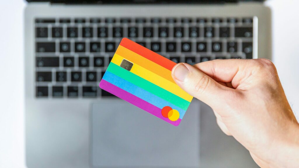 A hand holding a rainbow coloured credit card in front of a laptop computer. 